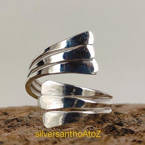 Könnte beinhalten: Ein silberfarbener Wickelring mit einem einzigartigen, geschichteten Design. Der Ring hat mehrere Bänder, die sich um den Finger krümmen und ein strukturiertes und auffälliges Aussehen erzeugen. Der Ring wird auf einer Holzoberfläche mit dem Text "silversanthoAtoZ" präsentiert.