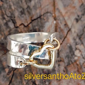 May include: A silver ring with a gold heart charm. The ring has a double band design and a gold heart pendant that hangs from the ring. The ring is on a brown and grey textured surface. The text "silversanthoAtoZ" is in the image.