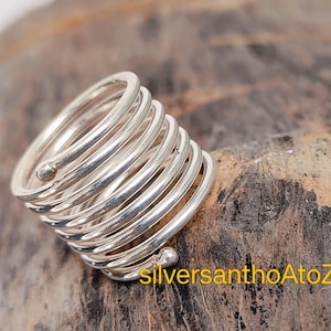 Puede incluir: Un anillo plateado en espiral con un diseño enrollado único. El anillo, hecho de metal brillante, presenta una serie de espirales redondeadas que crean una apariencia texturizada. Se muestra sobre una superficie marrón texturizada. El diseño es llamativo.