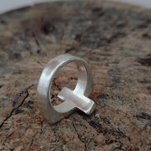 Puede incluir: Un anillo plateado con la forma de la letra "Q". El anillo est&aacute; hecho de un material met&aacute;lico brillante y se encuentra sobre un fondo marr&oacute;n texturizado. El dise&ntilde;o del anillo es simple y moderno.