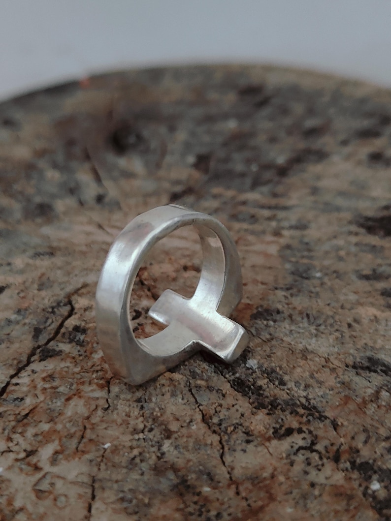 Puede incluir: Un anillo plateado con la forma de la letra "Q". El anillo est&aacute; hecho de un material met&aacute;lico brillante y se encuentra sobre un fondo marr&oacute;n texturizado. El dise&ntilde;o del anillo es simple y moderno.