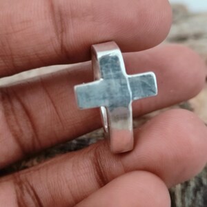 Puede incluir: Un anillo plateado con un dise&ntilde;o de cruz. La cruz est&aacute; centrada en el anillo y tiene una forma geom&eacute;trica simple. El anillo est&aacute; hecho de un metal brillante y parece estar bien elaborado.