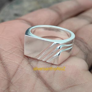 Könnte beinhalten: Nahaufnahme eines silbernen Rings mit quadratischer Oberfläche und diagonalen, geätzten Linien. Der Ring ist glänzend und reflektierend, mit dem Text "silversanthoAtoZ" in Gelb unten.