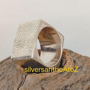 Op de afbeelding: Een zilverkleurige, vierkante ring met een getextureerd oppervlak aan de bovenkant. De ring heeft een geometrisch ontwerp en wordt gepresenteerd op een stuk hout. De binnenkant van de ring is glad en gepolijst.
