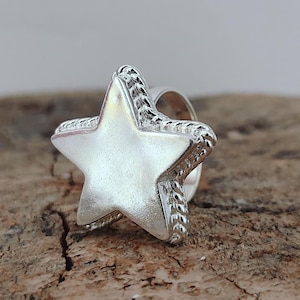 Puede incluir: Un anillo de estrella plateado. La estrella tiene un borde texturizado y un centro liso y pulido. El anillo está sobre una superficie de madera natural.