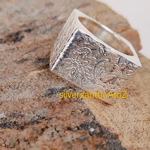 May include: A square silver ring with an ornate floral pattern. The ring has a textured surface and a polished interior. The text "silversanthoAtoZ" is visible on the surface. The ring is displayed on a brown, textured surface.