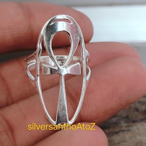 Peut inclure: Une bague argentée représentant un symbole Ankh, une croix avec une boucle en haut. La bague a un design ouvert, laissant apparaître les détails de l'Ankh. Le texte "silversanthoAtoZ" est imprimé en jaune en bas.