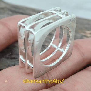 Pode incluir: Um conjunto de três anéis quadrados de prata, interligados e polidos. Os anéis são segurados em uma mão. O texto "silversanthoAtoZ" é visível na parte inferior da imagem. Os anéis são feitos de metal brilhante.