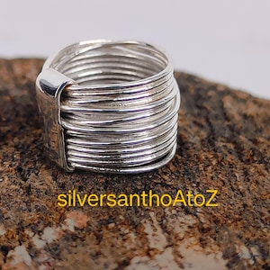 May include: A silver ring with a distinctive design. The ring comprises multiple bands of silver wire wrapped around the finger, with a rectangular accent. The ring is displayed on a textured, brown surface. The text "silversanthoAtoZ" is visible.