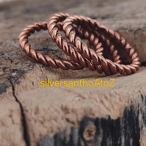 Op de afbeelding: Close-up van twee koperkleurige, gedraaide ringen in touwstijl. De ringen liggen op een verweerd houten oppervlak. De ringen staan in focus, met de achtergrond licht wazig. De tekst "silversanthoAtoZ" is zichtbaar op het hout.