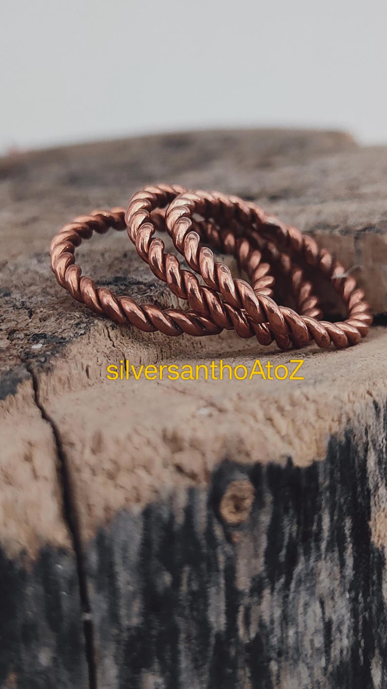 Op de afbeelding: Close-up van twee koperkleurige, gedraaide ringen in touwstijl. De ringen liggen op een verweerd houten oppervlak. De ringen staan in focus, met de achtergrond licht wazig. De tekst "silversanthoAtoZ" is zichtbaar op het hout.