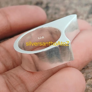 Puede incluir: Un anillo de plata con un diseño angular único. El anillo presenta una parte superior plana y rectangular que se extiende desde la banda. El texto "silversanthoAtoZ" es visible en el anillo. El anillo está hecho de un material metálico brillante.