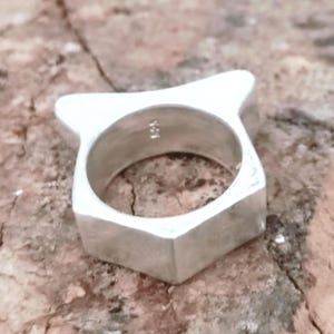 Puede incluir: Un anillo de plata con un diseño geométrico. El anillo tiene forma cuadrada con una parte superior puntiaguda y un acabado liso y pulido. El anillo se muestra sobre una superficie marrón texturizada.