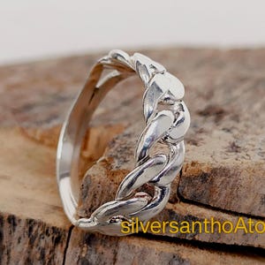 May include: A silver ring with a chain link design. The ring is made of shiny metal and has a braided appearance. The ring is displayed on a textured, brown surface.
