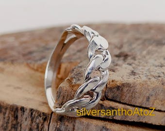 Könnte beinhalten: Ein silberner Ring mit einem Kettenglied-Design. Der Ring aus glänzendem Metall hat ein geflochtenes Aussehen. Er wird auf einer strukturierten, braunen Oberfläche präsentiert.