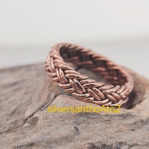 May include: A copper-coloured ring with a braided design. The ring is made of interwoven strands, creating a textured surface. The ring is displayed on a piece of wood.