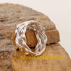 May include: A silver ring with a woven design. The ring is made of shiny silver metal and features a detailed braided pattern. The ring is displayed on a piece of wood.