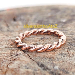 May include: A copper-colored ring with a twisted rope design. The ring is set against a rustic wooden surface. The text "silversanthoAtoZ" is visible in the image.