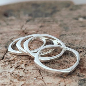 Op de afbeelding: Een verzameling van vier zilveren ringen met een geometrisch, vierkantachtig ontwerp. De ringen zijn gestapeld op een getextureerd, bruin houten oppervlak. De ringen hebben een gepolijste afwerking.