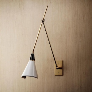 Puede incluir: Una lámpara de pared con una pantalla blanca en forma de cono y un acento negro. La lámpara presenta un brazo de color latón y una placa de pared rectangular. El diseño es moderno y minimalista.