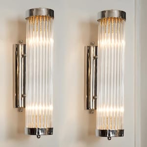 Könnte beinhalten: Zwei passende Wandleuchten mit zylindrischem Design. Jede Leuchte hat eine klare, geriffelte Glasröhre, die von einem polierten silberfarbenen Metallrahmen umschlossen ist. Die Ober- und Unterseite der Leuchten sind silberfarben, und das Innere strahlt golden.