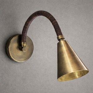 May include: A wall-mounted lamp featuring a circular brass backplate, a flexible arm wrapped in dark brown leather-like material, and a conical brass shade. The fixture has a metallic finish and a curved design that directs light downward.