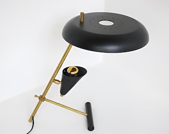 May include: A mid-century modern desk lamp with a black shade and a gold-colored, angled arm. The lamp features a cone-shaped light fixture and a round base. The lamp is made of metal and has a sleek, minimalist design.