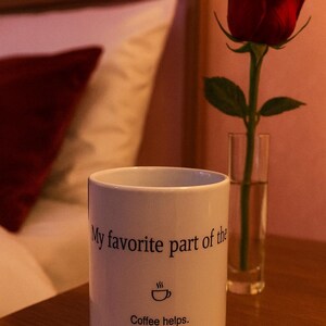May include: A white coffee mug with the text "My favourite part of the" and "Coffee helps." A red rose in a clear glass vase sits next to the mug. The mug has a coffee cup icon. The background is a bedroom setting.