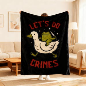 May include: Black blanket with a whimsical design featuring a white duck carrying a green frog holding a knife. The text "LET'S DO CRIMES" is printed in red. The blanket is held up by a person.