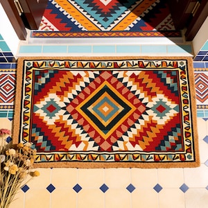 Puede incluir: Una alfombra rectangular con un patrón geométrico en rojo, naranja, amarillo, azul y blanco. El diseño central es un rombo con otro rombo más pequeño en su interior. La alfombra tiene un borde marrón y está colocada sobre un suelo embaldosado. Mide aprox. 90 cm x 60 cm.
