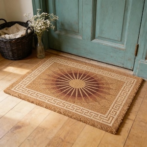 May include: Rectangular doormat with a sunburst design in shades of brown and tan. The sunburst is centered within a Greek key border. The mat is made of a natural fiber material and is placed in front of a teal door.
