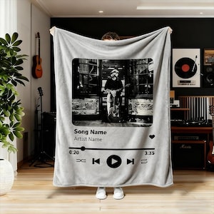 May include: A light gray blanket featuring a black and white image of a musician with a guitar. The blanket design resembles a music player interface, with text reading "Song Name" and "Artist Name". The blanket is held up by a person.