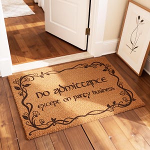 May include: A rectangular doormat with the text "no admittance except on party business" in black script. The mat is brown with a decorative black floral border. The mat is placed in front of a white door on a hardwood floor.