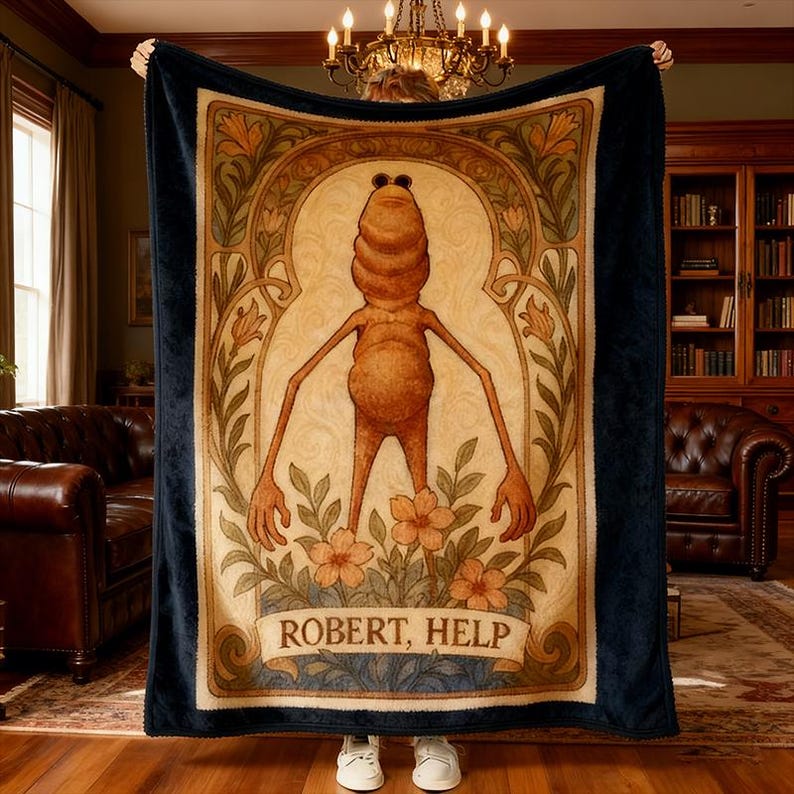 Puede incluir: Una manta azul marino con una ilustraci&oacute;n central de una figura estilizada y el texto "ROBERT, HELP". La obra de arte est&aacute; enmarcada por elementos florales y arqueados en tonos beige, marr&oacute;n y verde. La manta est&aacute; sostenida por una persona que lleva zapatillas blancas.