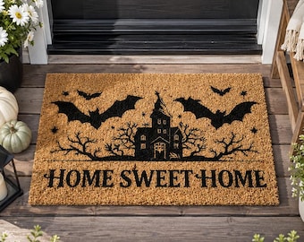May include: A rectangular tan coir doormat featuring a black graphic print of a haunted house, bare trees, and flying bats. The text "HOME SWEET HOME" is printed in a bold, black serif font across the bottom portion of the mat.