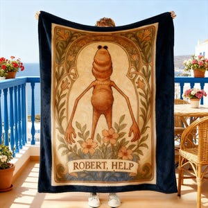Puede incluir: Una manta azul marino con una ilustraci&oacute;n central de una figura antropom&oacute;rfica estilizada con gafas de sol, rodeada de elementos florales y de inspiraci&oacute;n art nouveau. El texto "ROBERT, HELP" se muestra en la parte inferior. La manta est&aacute; sujeta por una persona.