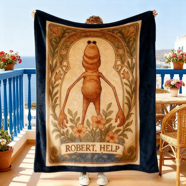 Puede incluir: Una manta azul marino con una ilustraci&oacute;n central de una figura antropom&oacute;rfica estilizada con gafas de sol, rodeada de elementos florales y de inspiraci&oacute;n art nouveau. El texto "ROBERT, HELP" se muestra en la parte inferior. La manta est&aacute; sujeta por una persona.