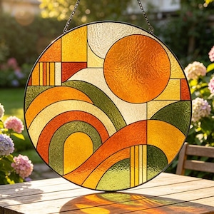 Può includere: Una finestra rotonda in vetro colorato con un design geometrico. La finestra presenta un grande cerchio arancione, con forme curve arancioni, gialle e verdi. La finestra è appesa a una catena e ha un bordo nero.
