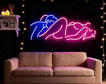 Pode incluir: Uma placa de néon que retrata um casal em um abraço romântico, com iluminação azul e rosa. A placa está montada em uma parede preta acima de um sofá bege. Luzes de corda e velas criam um ambiente acolhedor.