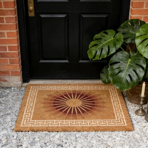 May include: A rectangular doormat with a geometric sunburst design in shades of brown and burgundy. The mat has a Greek key border and is made of natural fibers. Placed in front of a black door, it adds a welcoming touch to any entryway.