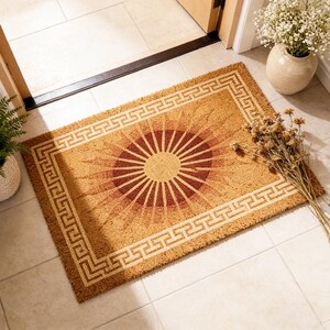 May include: Rectangular doormat with a sunburst design in shades of brown and tan. The center features a dark red circle with radiating lines, surrounded by a lighter brown gradient. A Greek key border frames the design, adding a touch of classic style.