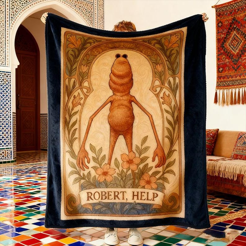 Puede incluir: Una manta rectangular grande con un borde azul oscuro. El centro presenta una ilustraci&oacute;n caprichosa de una figura humanoide marr&oacute;n con el texto "ROBERT, HELP". El dise&ntilde;o est&aacute; enmarcado por elementos florales y art nouveau.
