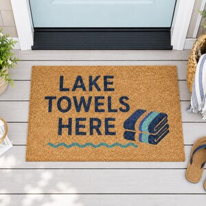 以下が含まれることがあります： 天然ココナッツ繊維で作られた長方形のドアマット。「LAKE TOWELS HERE」の文字がネイビーブルーで、積み重ねられたタオルと波線がネイビーとティールで描かれています。玄関ドアの前に置かれています。