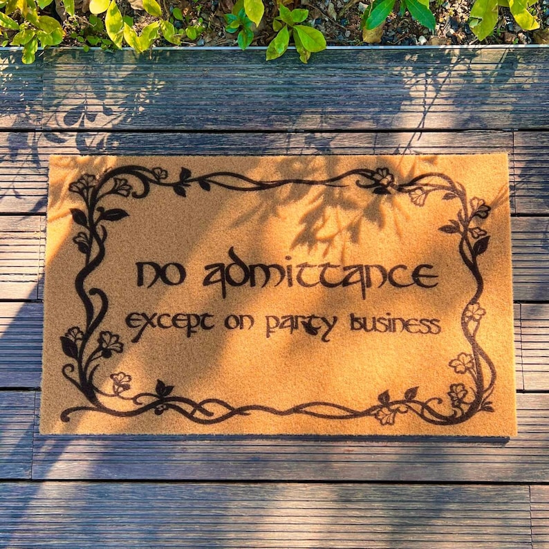May include: A rectangular doormat with the text "NO admittance except on party business" in black. The mat is tan with a black floral border. The mat is on a wooden deck.