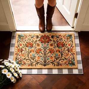 Op de afbeelding: Een rechthoekige deurmat met een bloemenpatroon in oranje, blauw en groen op een beige achtergrond. De mat ligt op een grijs-wit geruit tapijt. Bruine laarzen zijn zichtbaar in de deuropening.