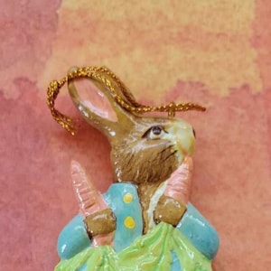 May include: A ceramic bunny figurine, wearing a blue jacket and holding carrots and green leaves. The bunny has brown fur, pink ears, and a gold string for hanging. A charming Easter decoration.