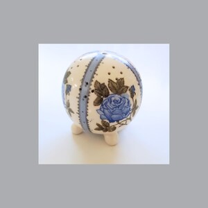 Könnte beinhalten: Ein weißer Keramik-Nähkissen in Kugelform mit drei kleinen Füßen. Das Nähkissen ist mit einem blauen und grünen Blumendesign verziert, das eine große Rose zeigt.