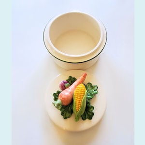 Puede incluir: Una caja de cerámica blanca con un borde verde y una tapa con un diseño 3D de una zanahoria, maíz y un nabo sobre un lecho de hojas verdes.