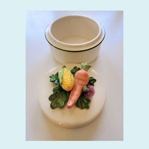 May include: White ceramic trinket box with a green trim. The lid features a 3D design of a carrot, corn, and a purple beetroot on a bed of green leaves.