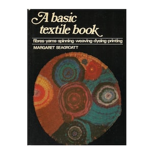 May include: A basic textile book with a black cover. The title is in white script, with the subtitle listing fibers, yarns, spinning, weaving, dyeing, and printing. The cover features a colorful circular design with various concentric circles.